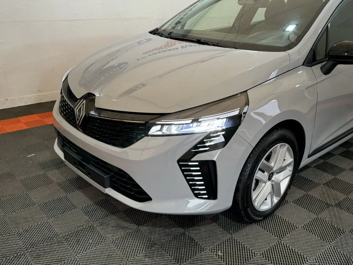 Vue 3/4 avant droite d'une Renault Clio gris clair 2025 avec phares LED allumés et jantes alliage modernes.