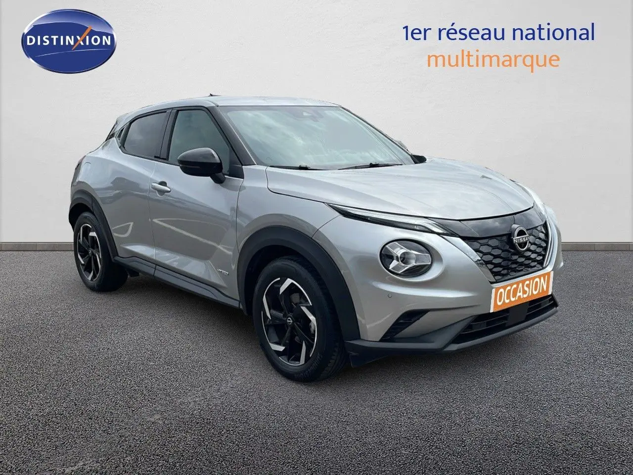 Nissan Juke hybride gris perle métal en 3/4 avant droit, avec jantes noires et calandre distinctive.