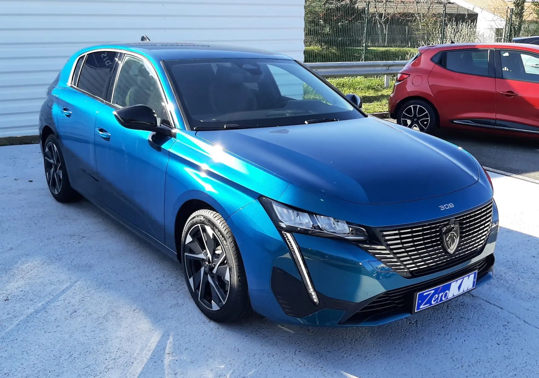 Vue frontale d'une Peugeot 308 Bleu Obsession 2026 avec calandre noire et logo lion distinctif, stationnée devant un mur blanc.