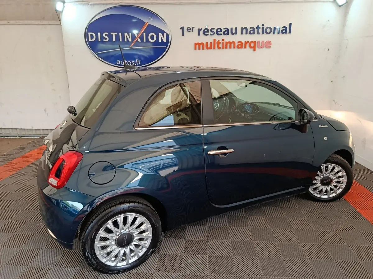 Vue latérale droite d'une FIAT 500 Dolcevita hybride bleu foncé avec jantes alliage et toit noir en intérieur showroom.
