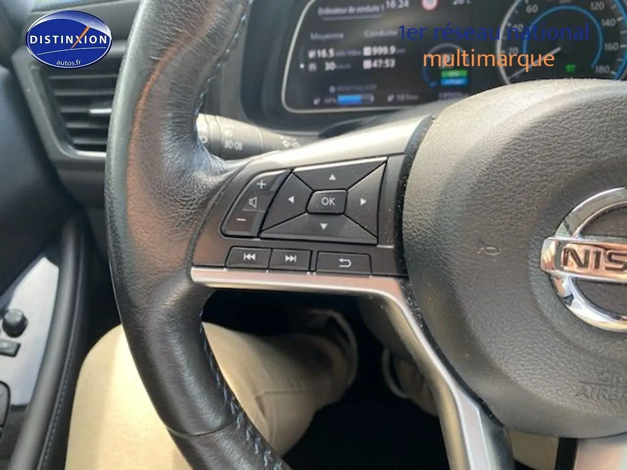 Gros plan sur le volant en cuir noir d'une Nissan Leaf électrique blanche nacrée, avec commandes intégrées et tableau de bord numérique visible.