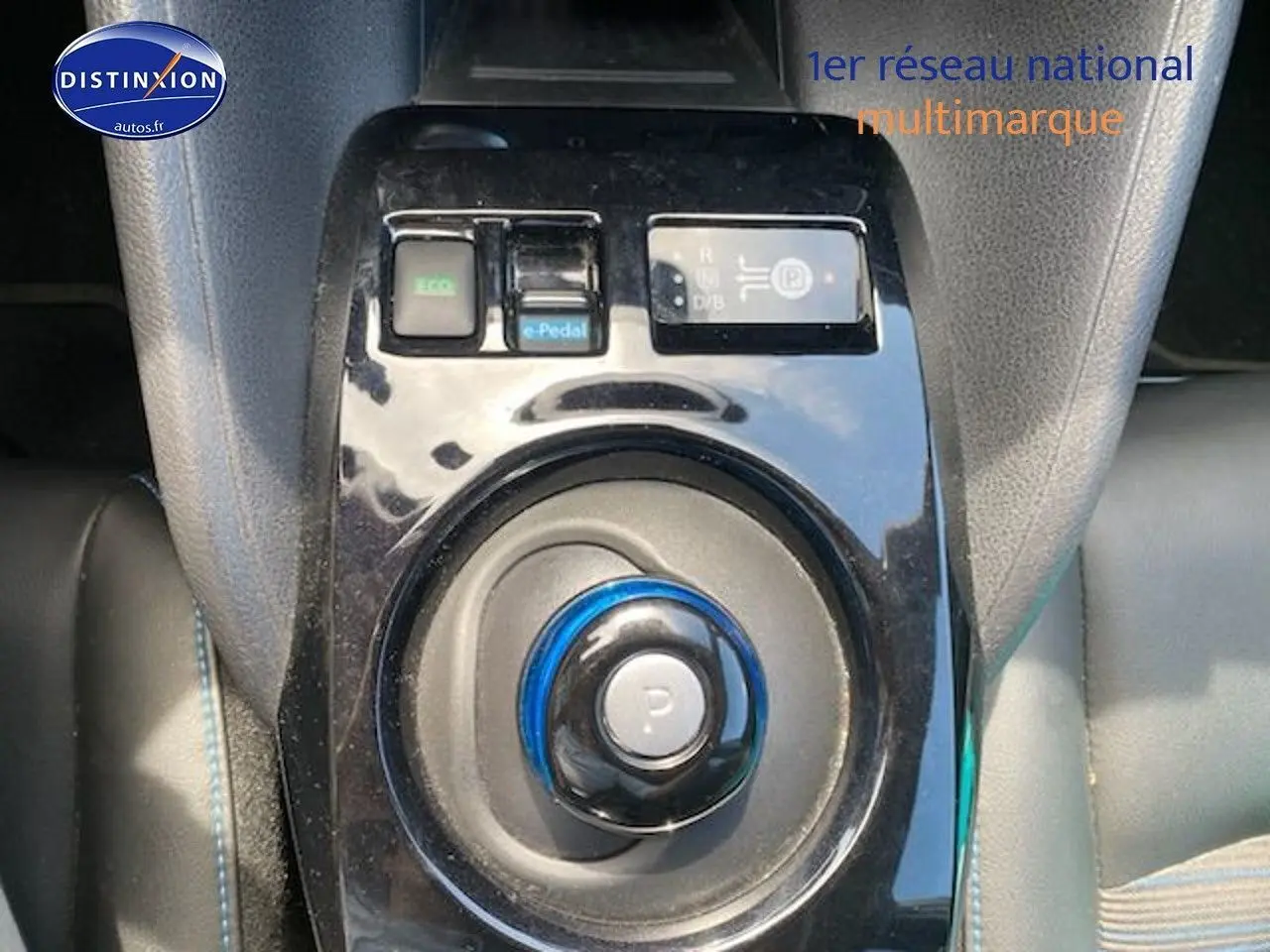 Gros plan sur la console centrale noire brillante avec sélecteur de vitesse rond et boutons de mode de conduite du Nissan Leaf blanc nacré.