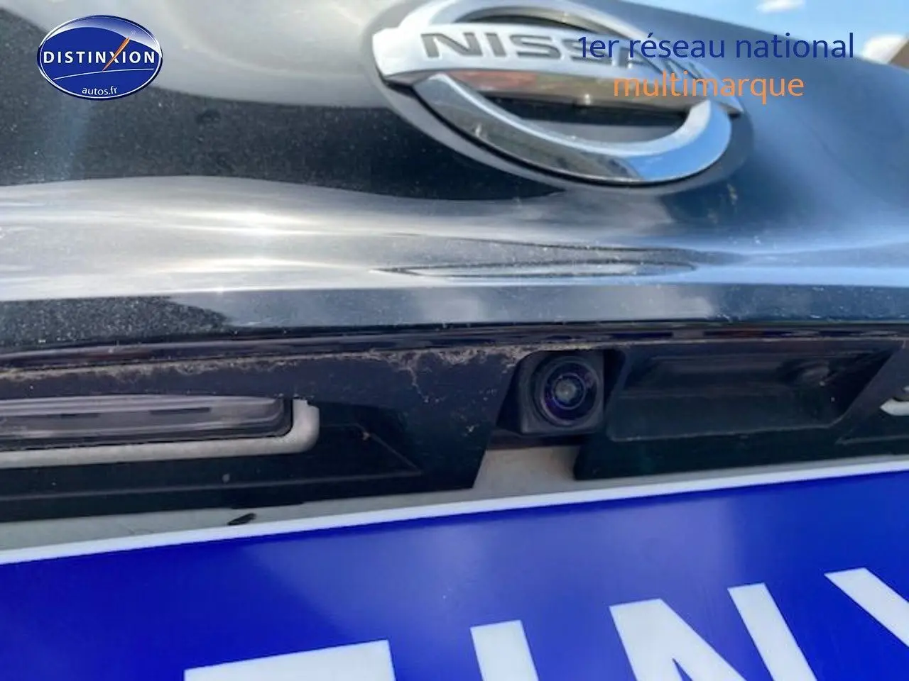 Gros plan sur la caméra de recul intégrée sous le logo Nissan d'une Leaf électrique blanc nacré.