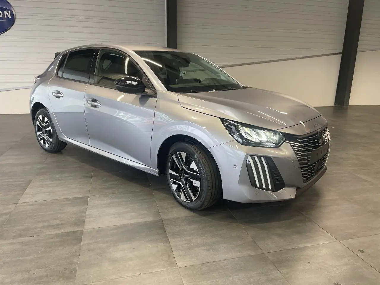 Peugeot 208 gris Artense vue 3/4 avant droit en intérieur avec jantes alliage bi-tons et calandre noire grainée.