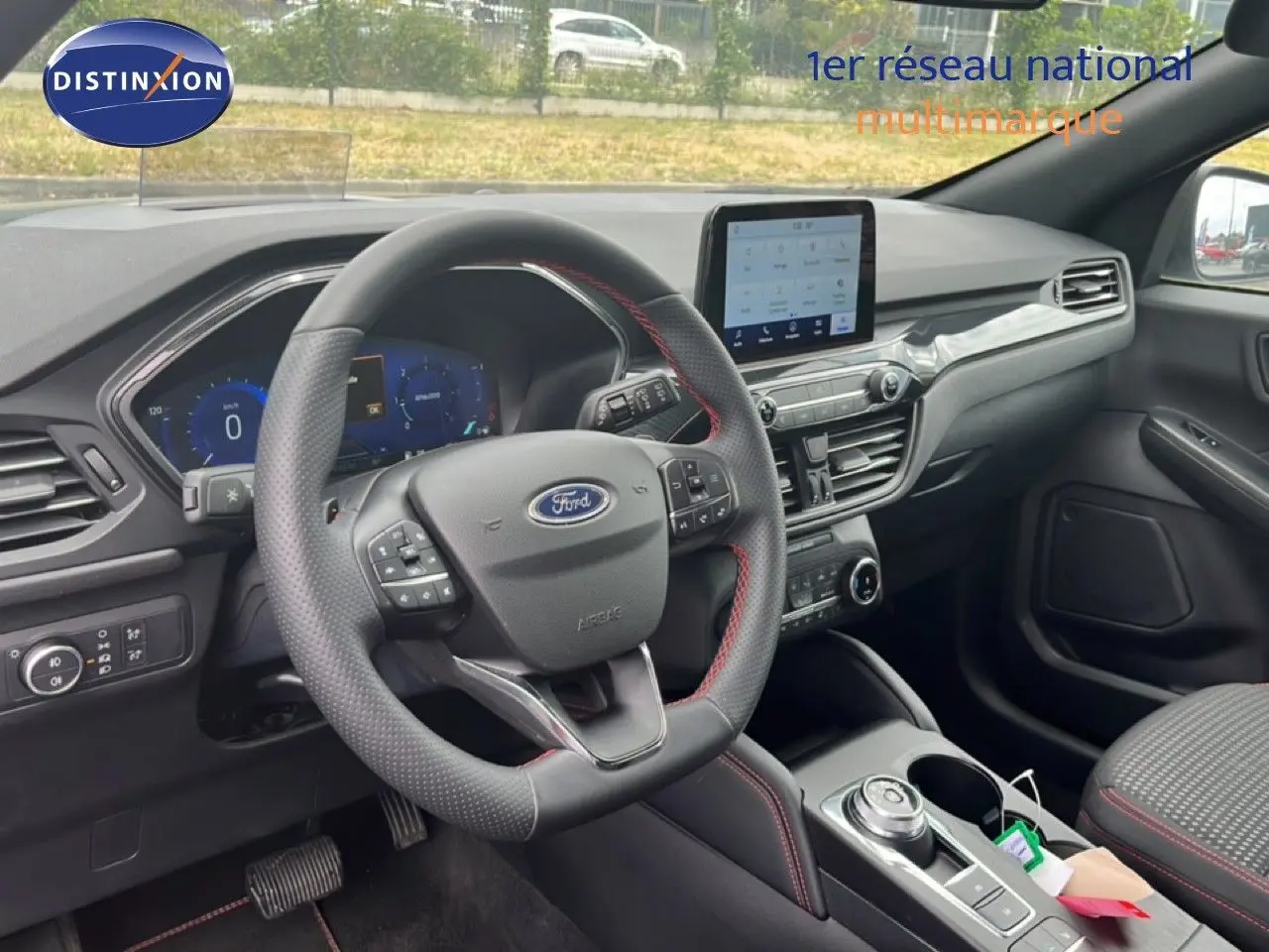 Vue intérieure côté conducteur du Ford Kuga 2024, tableau de bord moderne avec écran tactile et volant cuir noir à surpiqûres rouges.