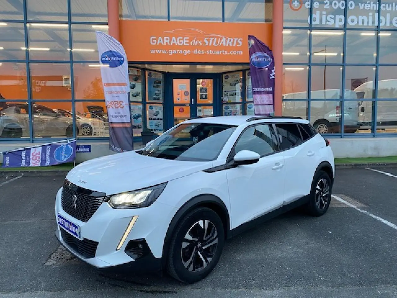 Peugeot 2008 blanc vue 3/4 avant droit, jantes alliage bi-ton et calandre noire devant un garage.