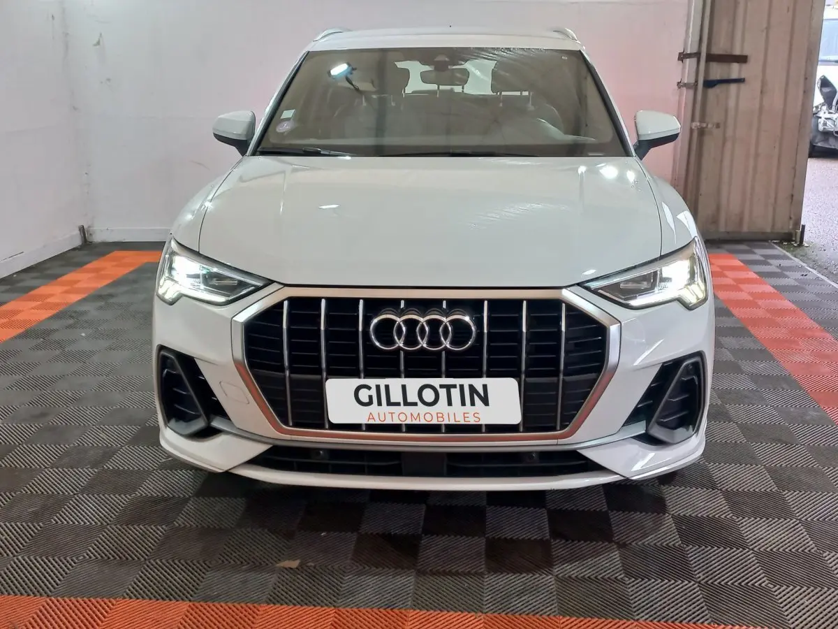 Audi Q3 blanc vue de face, avec calandre large à barres verticales et phares LED allumés dans un garage.