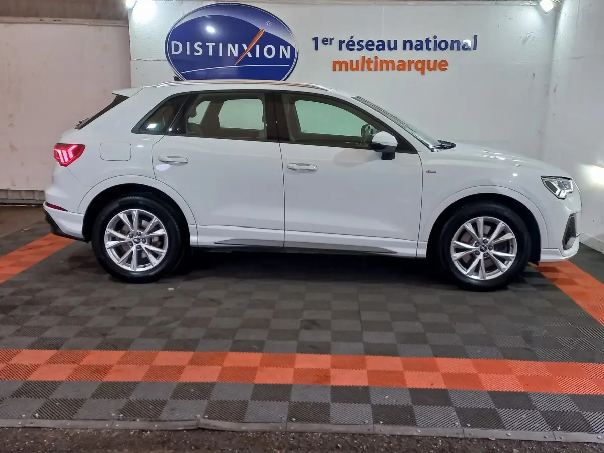 Audi Q3 blanc en vue de profil côté gauche, stationné en intérieur avec jantes alliage et feux arrière allumés.