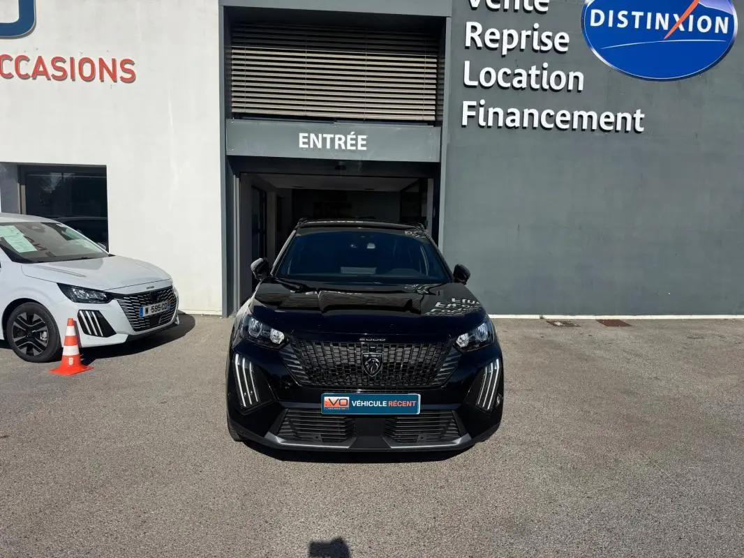 Peugeot 2008 Hybrid noir vue de face, avec barres de toit et calandre distinctive, stationné devant un bâtiment commercial.