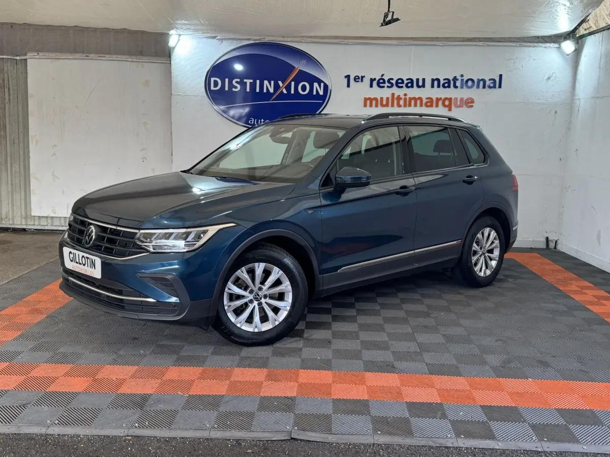 Volkswagen Tiguan bleu en 3/4 avant droit avec rails de toit noirs, stationné sur sol à damiers gris et orange.