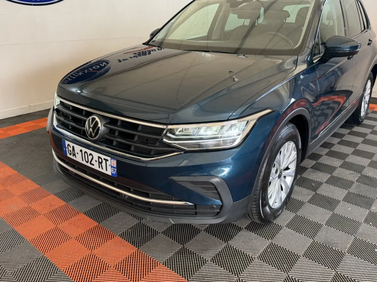 Volkswagen Tiguan bleu en 3/4 avant droit, avec calandre chromée et phares LED allumés, sur sol damier noir et orange.