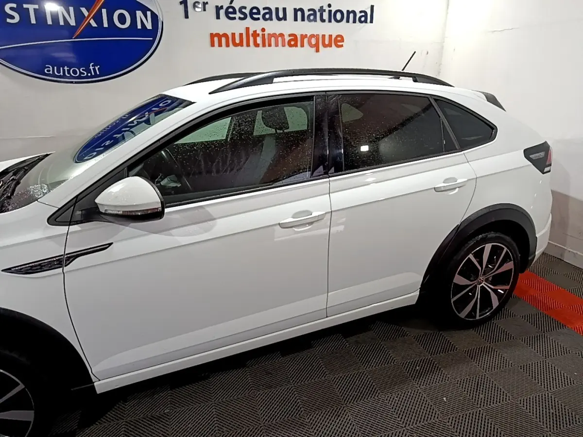 Vue de profil côté gauche d'un Volkswagen Taigo blanc avec jantes alliage et rails de toit noirs en intérieur showroom.