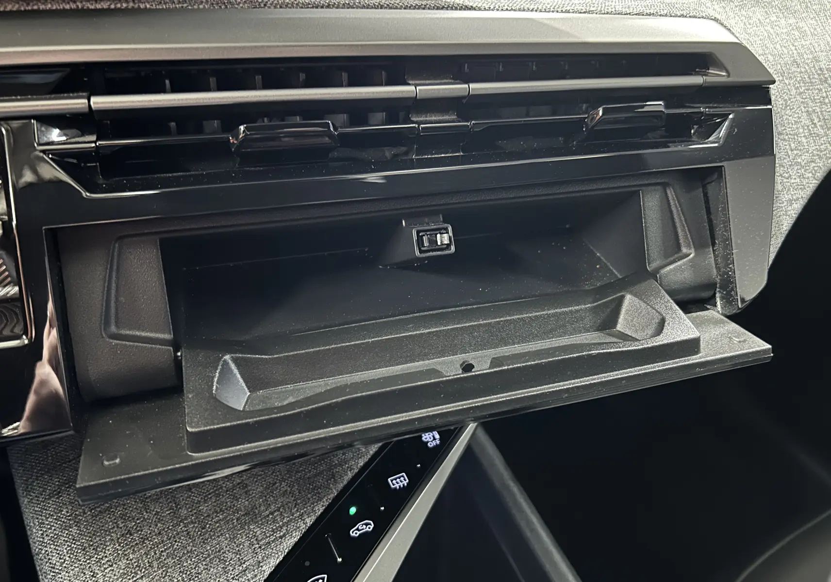 Gros plan sur la boîte à gants ouverte et la console centrale noire du Peugeot 5008 hybride 2025, intérieur gris.