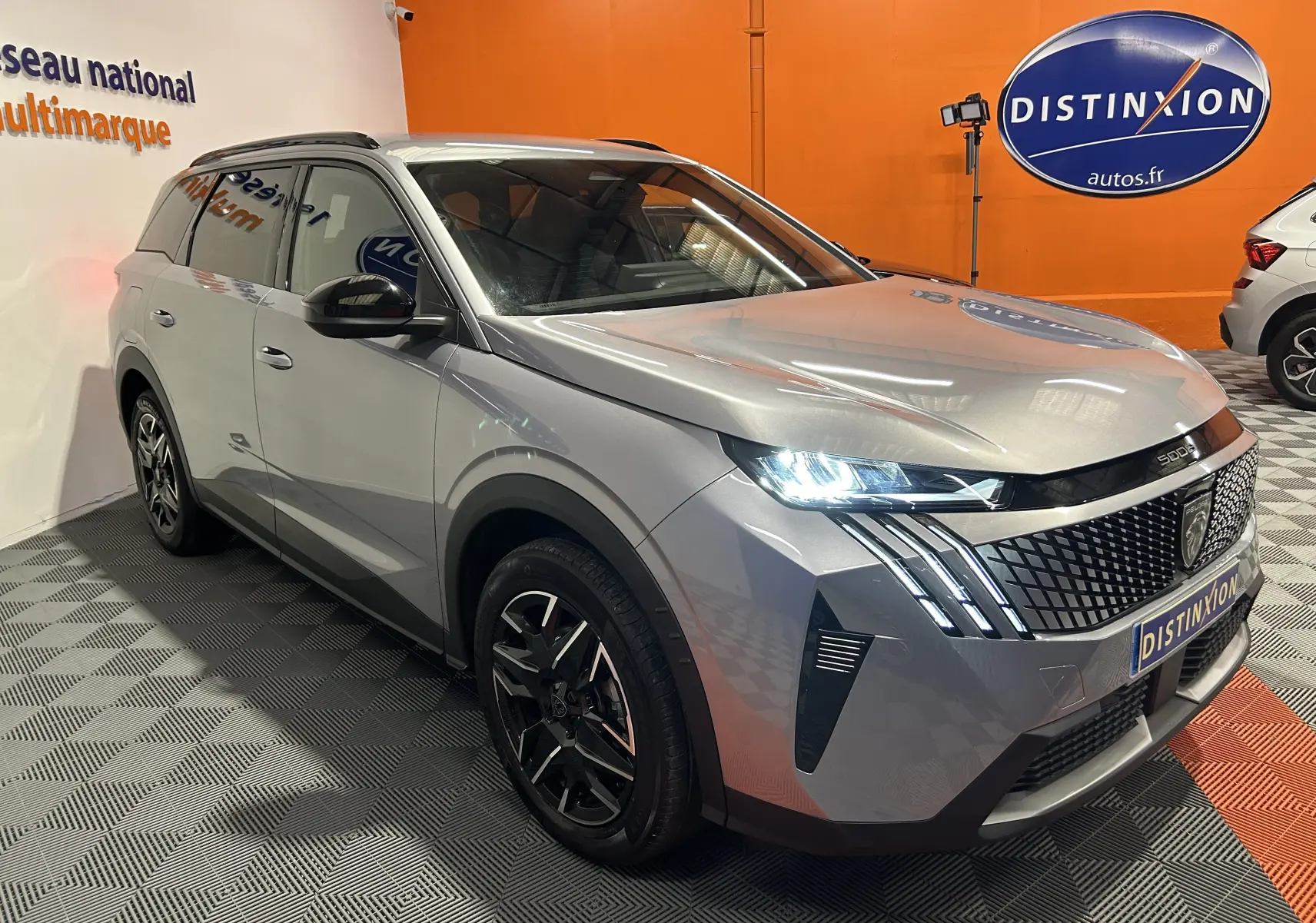 Peugeot 5008 gris Artense en 3/4 avant droit, avec phares LED allumés et calandre distinctive en showroom.