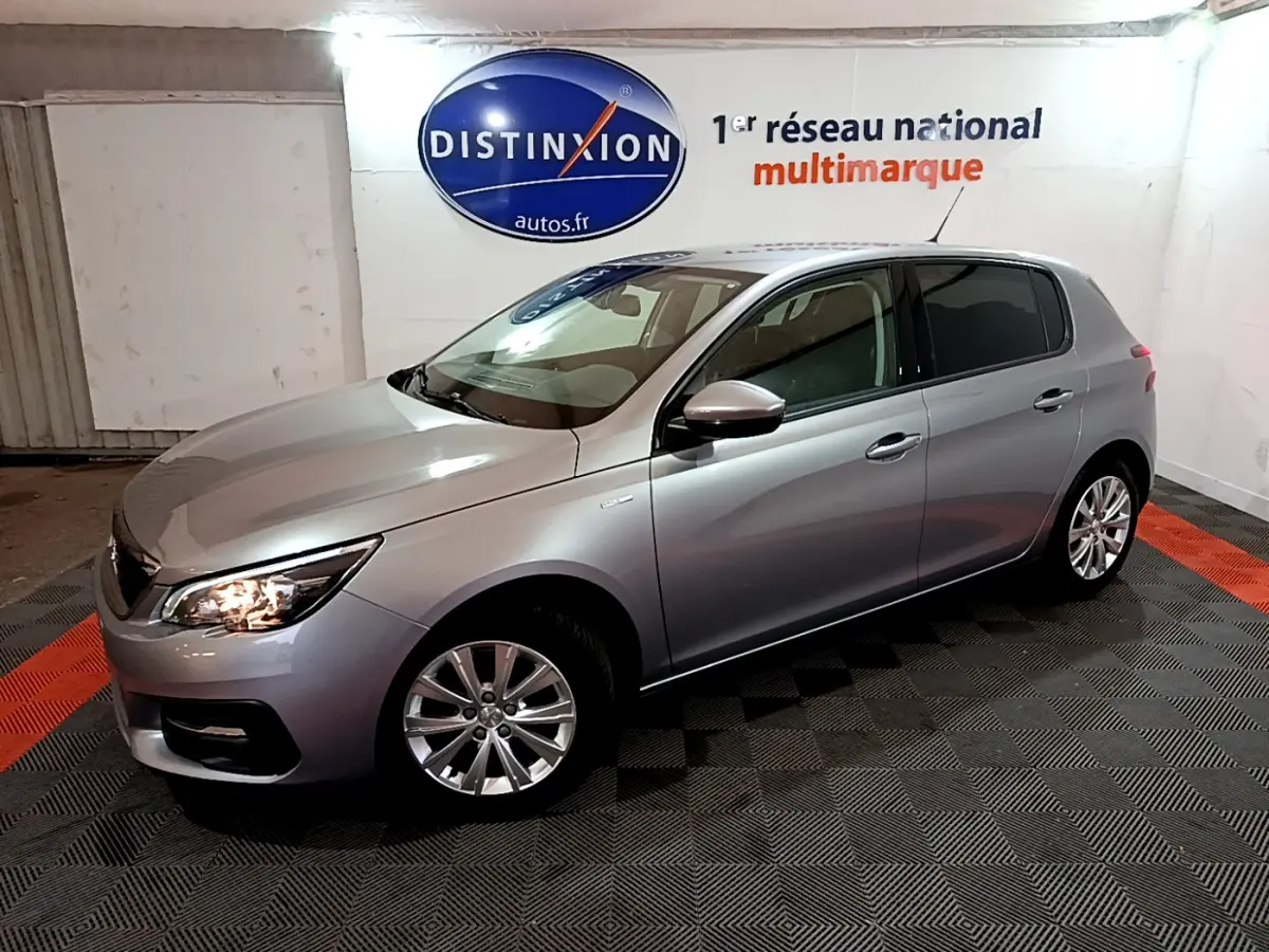 Peugeot 308 Puretech gris clair vue 3/4 avant droit, phares allumés, dans un showroom intérieur.