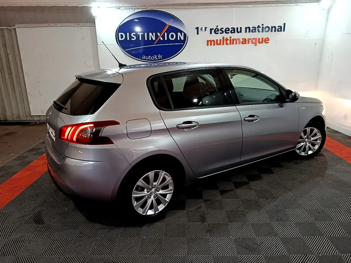 Peugeot 308 gris clair vue 3/4 arrière côté droit, feux arrière allumés dans un showroom intérieur.