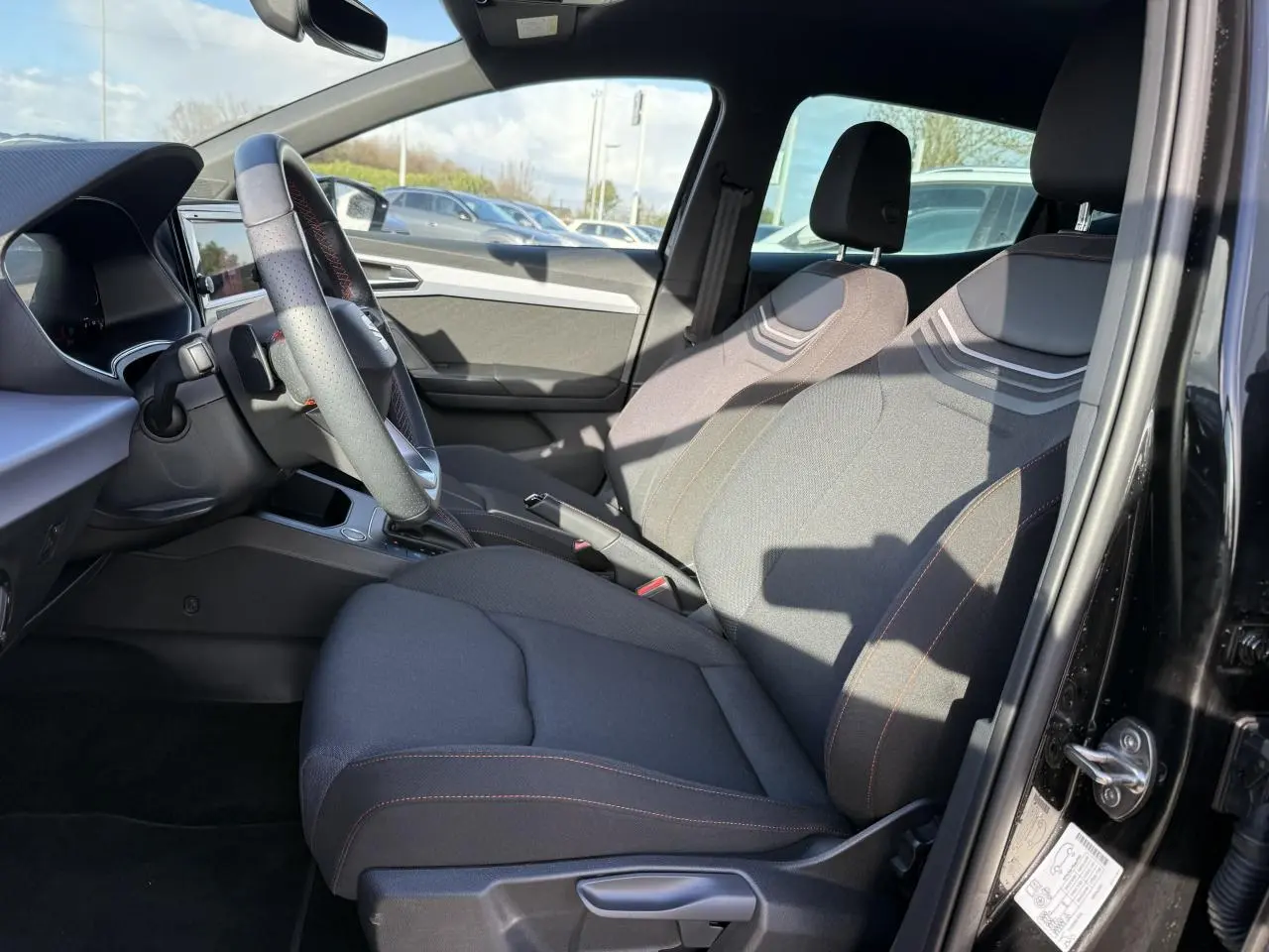 Intérieur de la SEAT IBIZA 2024 vue côté conducteur, sièges tissu gris foncé avec surpiqûres rouges et volant cuir multifonction.