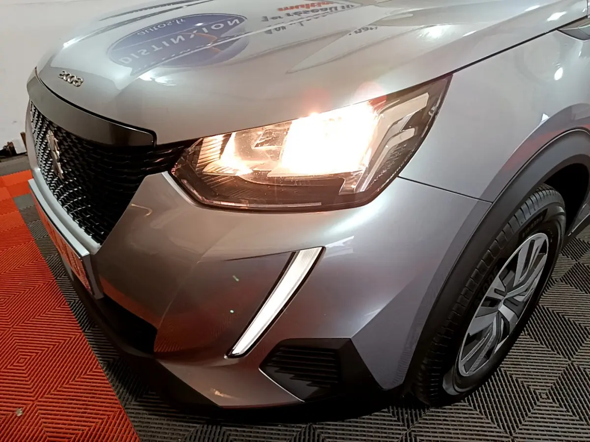 Gros plan sur l'avant droit d'un Peugeot 2008 gris clair 2022 avec phares allumés et calandre noire.