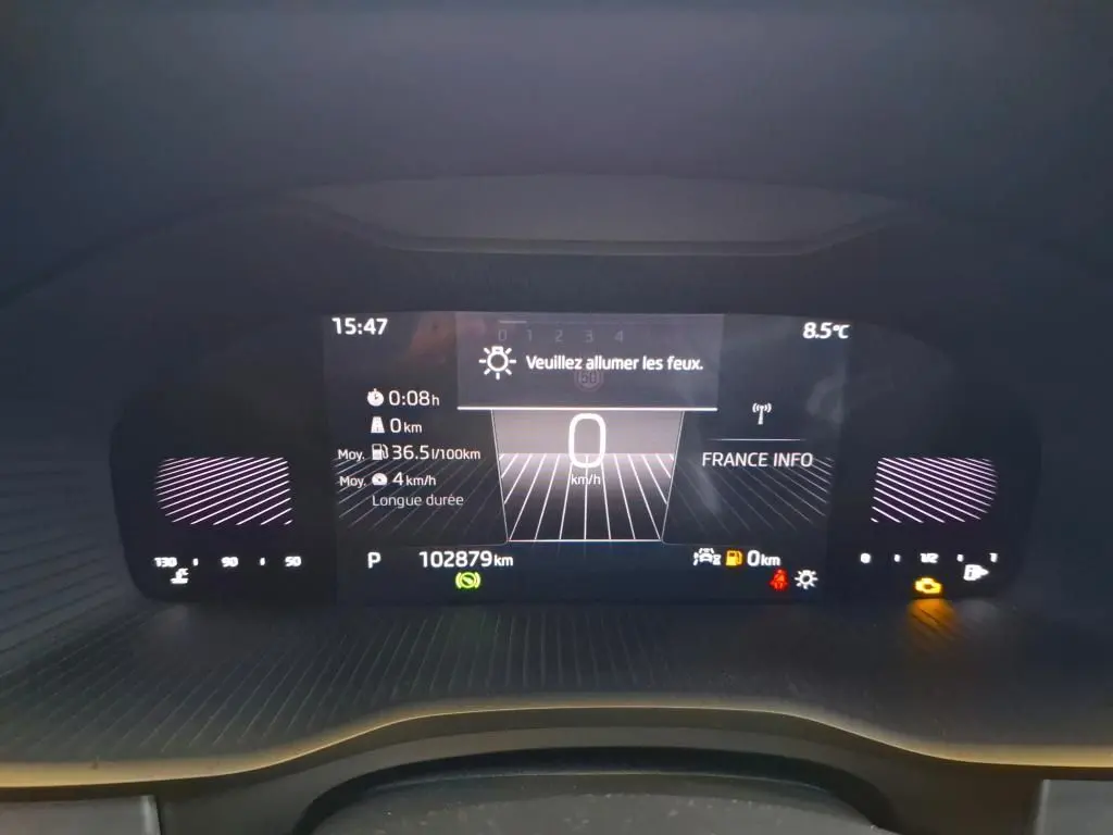 Tableau de bord numérique du Skoda Kodiaq gris clair, affichant la vitesse à 0 km/h et une alerte pour allumer les feux.