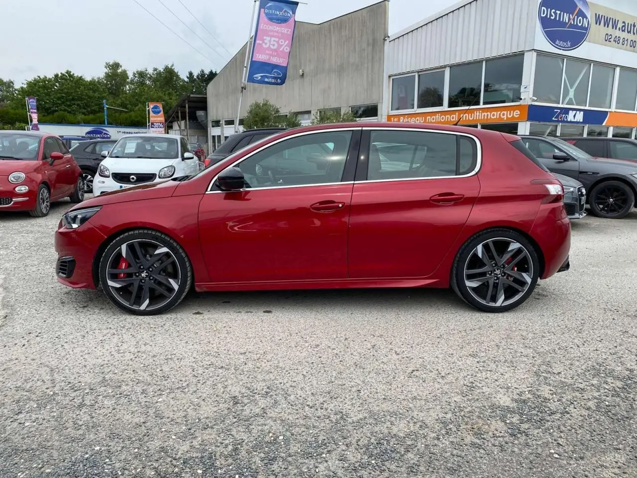 Profil latéral droit d'une Peugeot 308 GTi rouge métallisée garée devant un concessionnaire multimarque.