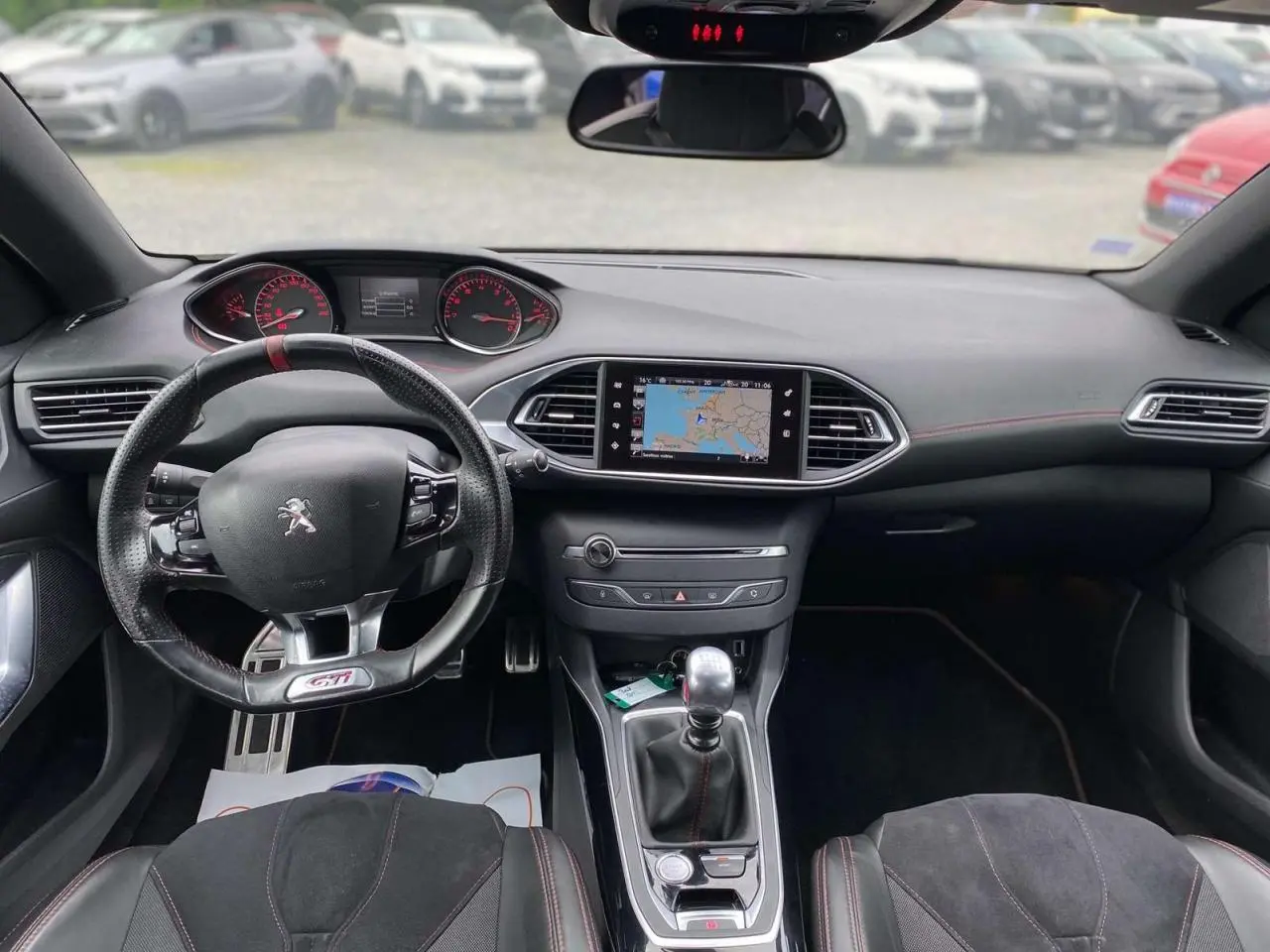 Vue intérieure avant du tableau de bord et volant GTi de la Peugeot 308 rouge, avec écran de navigation et boîte manuelle.