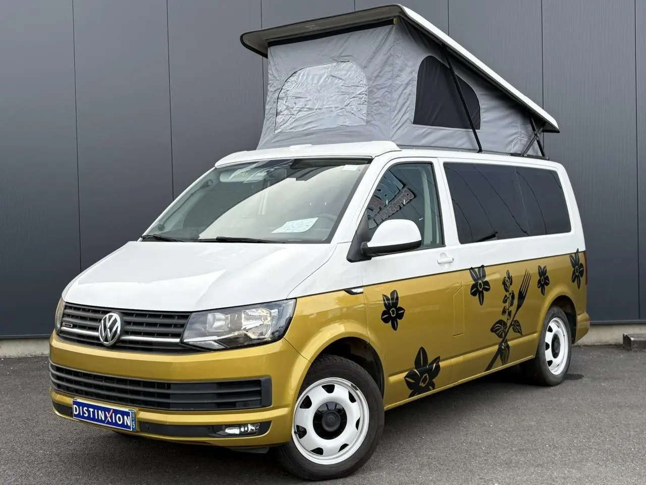 Vue 3/4 avant droite du Volkswagen Transporter aménagé jaune et blanc avec toit relevé et motifs floraux noirs.