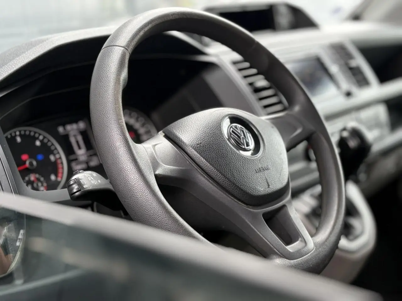 Vue rapprochée du volant noir du Volkswagen Transporter 2017, avec tableau de bord et commandes visibles en arrière-plan.