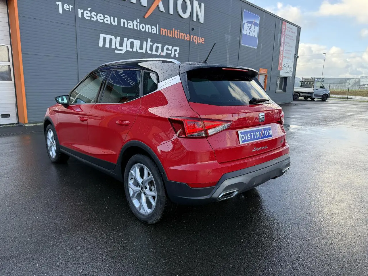 Vue 3/4 arrière droite d'un SEAT Arona Rouge Pur 2024 avec toit noir et jantes argentées sur sol mouillé.