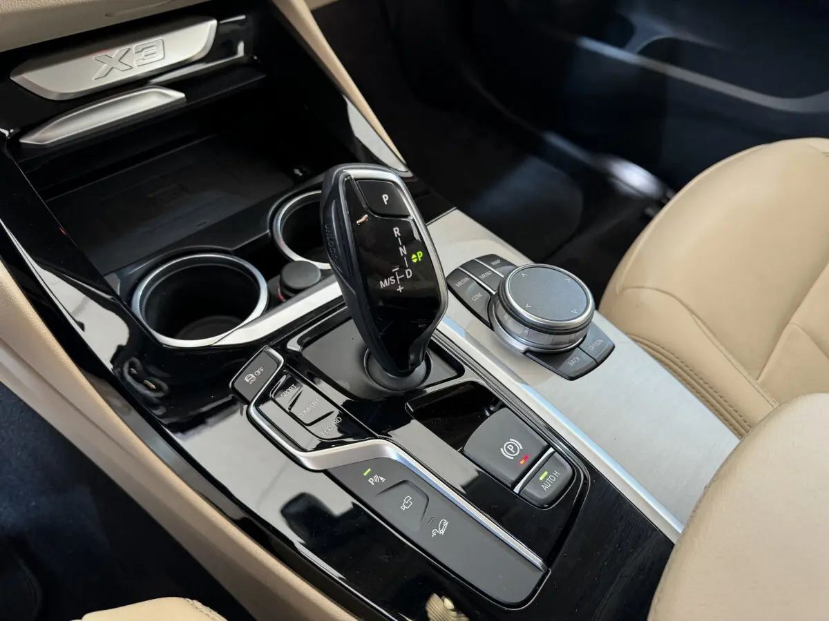 Vue rapprochée du levier de vitesse automatique et molette de commande sur console noire brillante d’un BMW X3 gris foncé, siège beige visible.