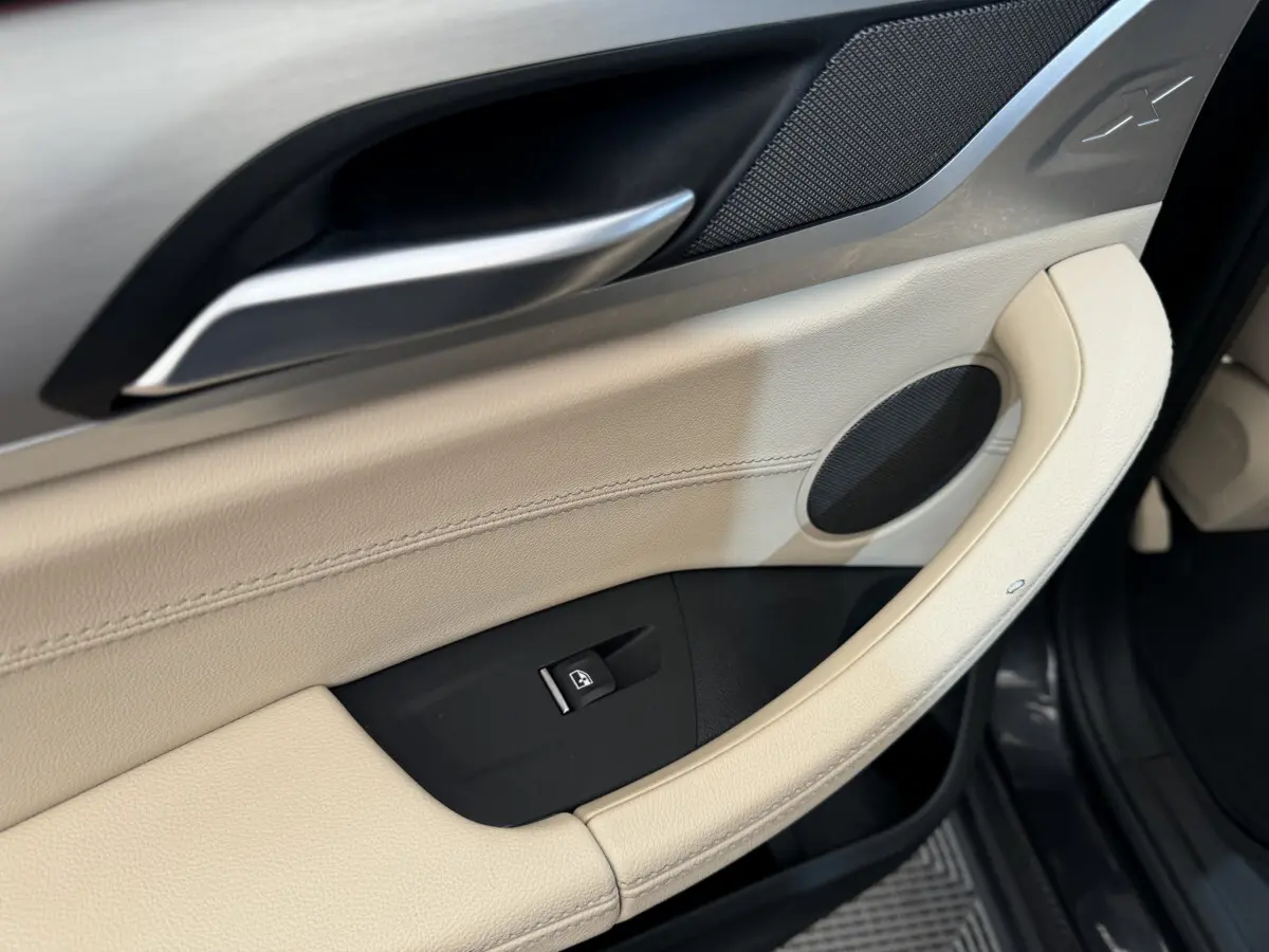 Gros plan sur la poignée intérieure beige et la commande de vitre du côté gauche d’un BMW X3 gris foncé.