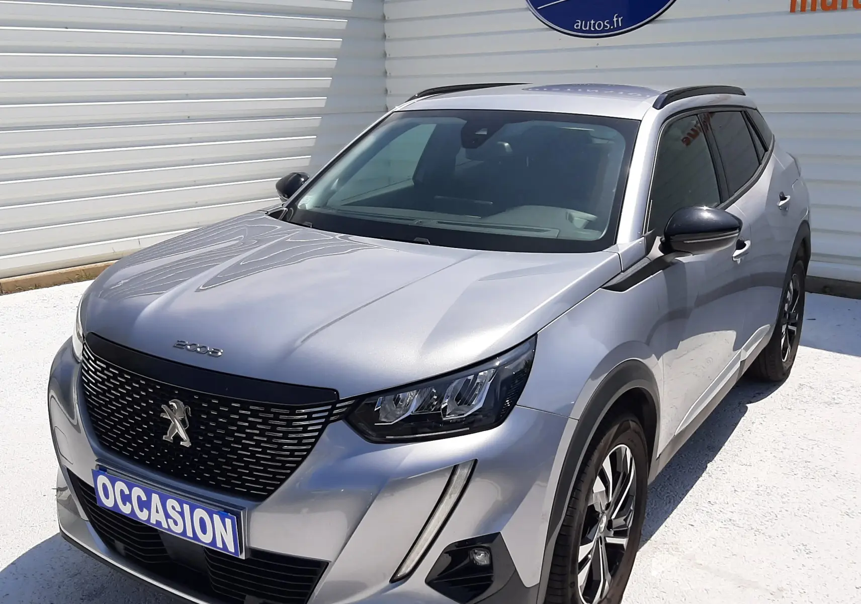 Peugeot 2008 gris Artense vue 3/4 avant droit, avec calandre noire et phares LED distinctifs, stationnée devant un mur blanc.