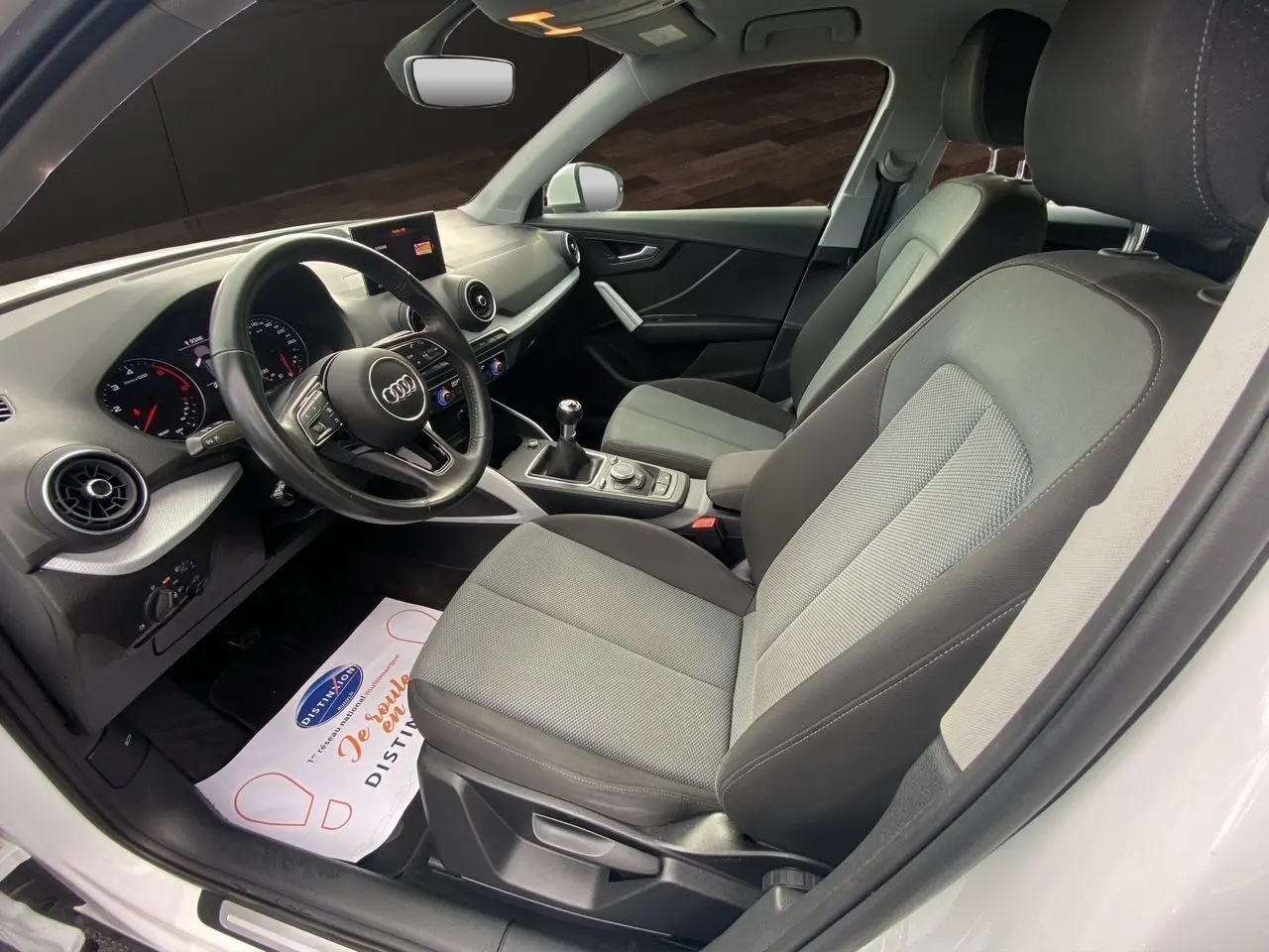 Vue intérieure côté conducteur de l'Audi Q2 30 TDI blanc, montrant les sièges tissu gris et le tableau de bord moderne.