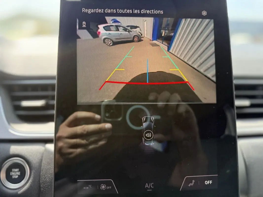 Vue écran caméra de recul montrant une Renault Captur gris Rafale avec toit noir, garée près d’un bâtiment industriel.