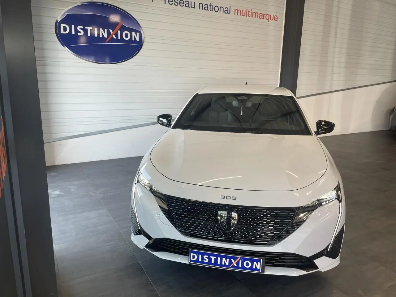 Vue avant d'une Peugeot 308 blanche 2025 avec calandre noire brillante et feux LED griffés, en showroom.