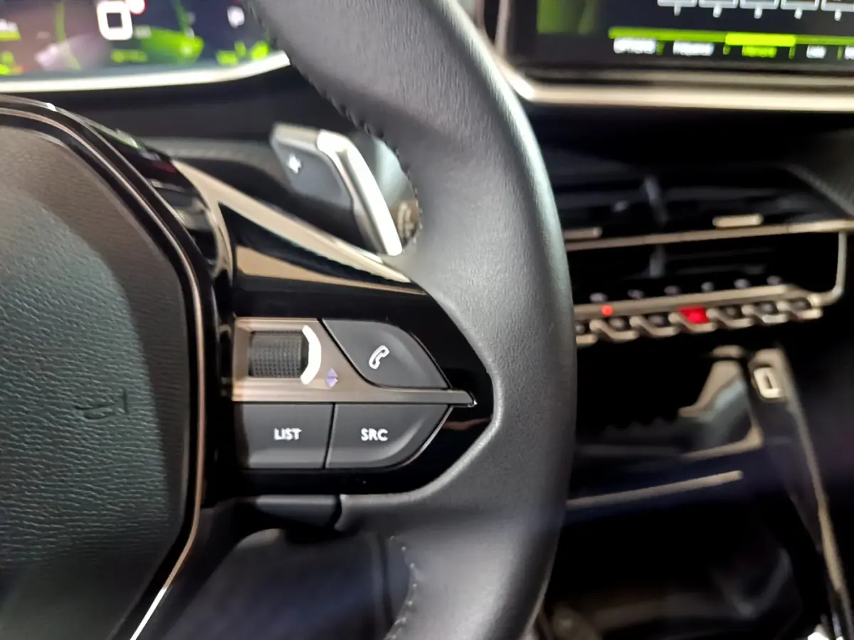 Gros plan sur les commandes gauche du volant cuir de la Peugeot 208 jaune 2023, avec tableau de bord numérique en arrière-plan.