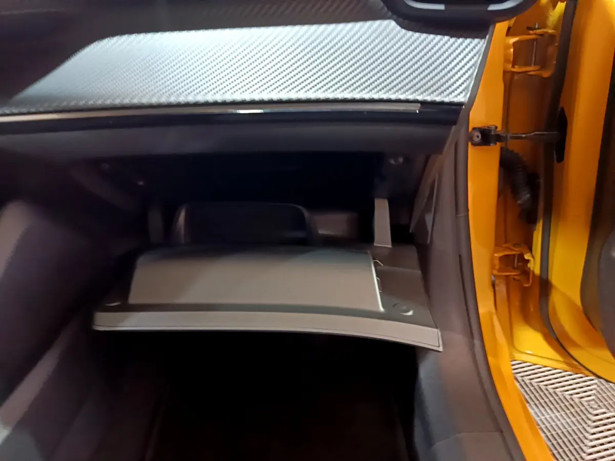 Gros plan sur la boîte à gants ouverte côté passager d'une Peugeot 208 jaune 2023, intérieur sombre et détails carbone.