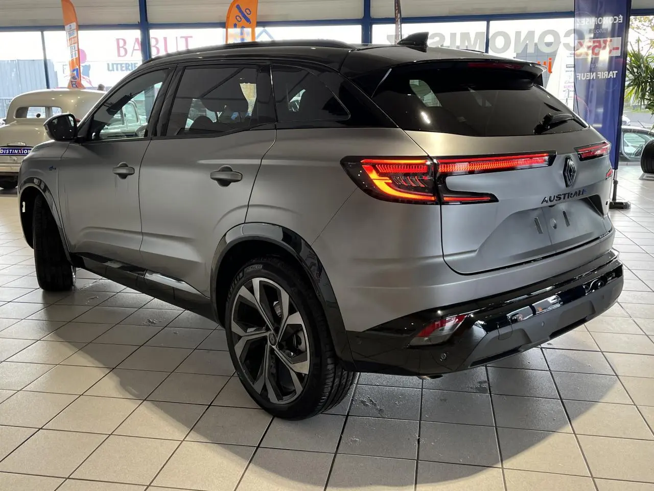 Vue 3/4 arrière droite du Renault Austral gris avec toit noir étoilé et feux arrière LED allumés en intérieur showroom.
