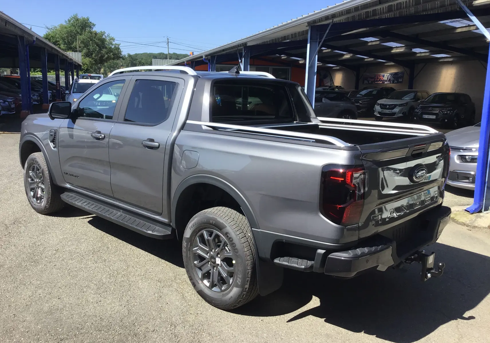 Ford Ranger Double Cabine gris carbone vu en 3/4 arrière droit, avec ridelle et attelage visibles sous un auvent.