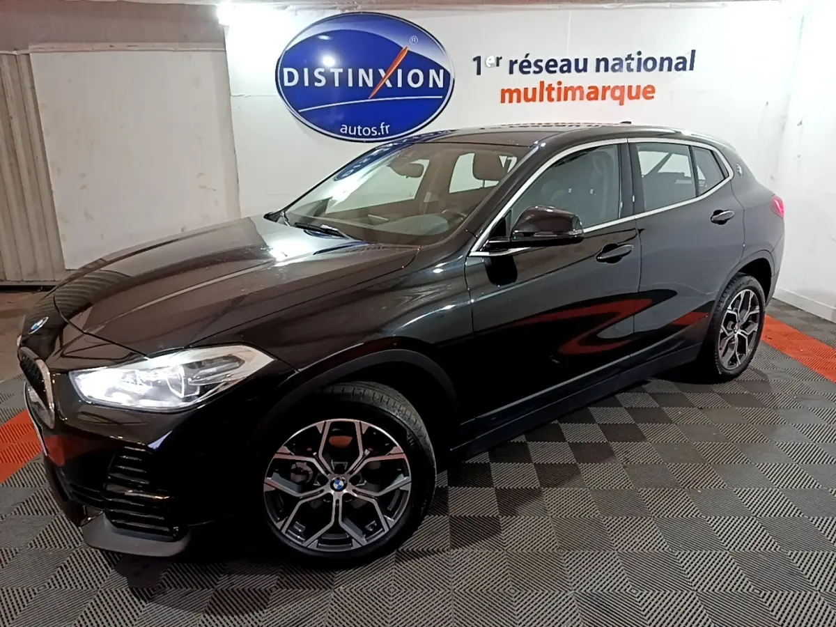 BMW X2 sDrive18i Lounge noir en 3/4 avant droit, avec jantes alliage et phares LED allumés dans un showroom.