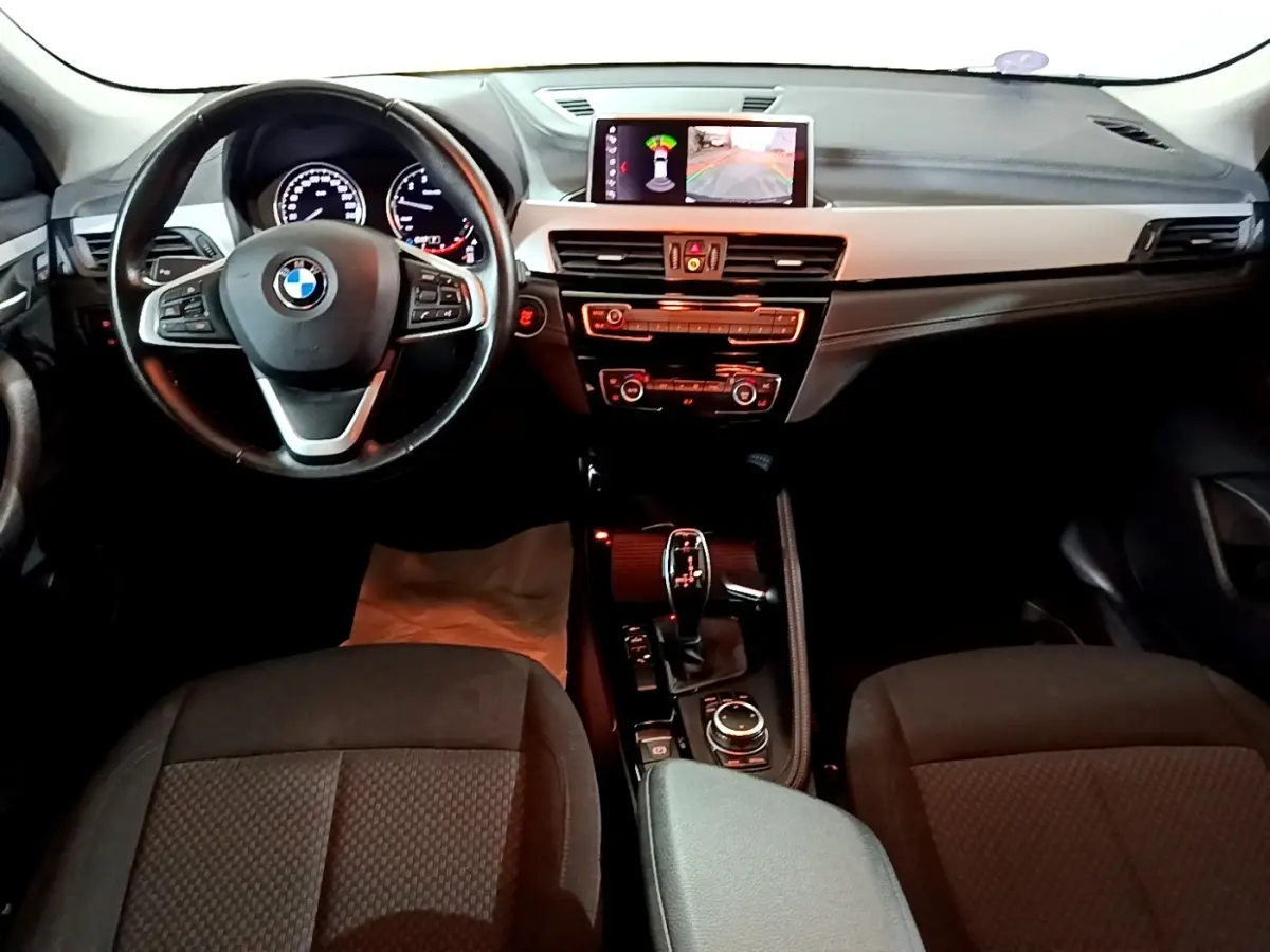 Vue intérieure avant du BMW X2 sDrive18i Lounge 2023, volant cuir alu multifonctions et écran central affichant l'aide au stationnement.