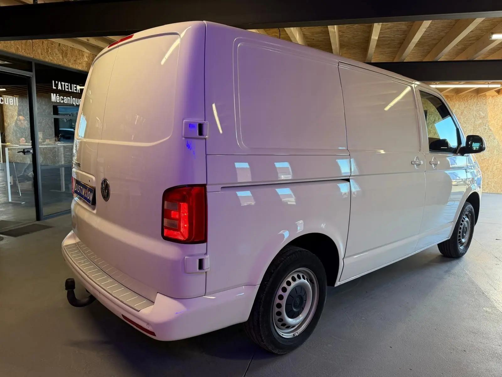 Vue 3/4 arrière droit d’un Volkswagen Transporter blanc 2019 en intérieur, avec feux arrière allumés et attelage visible.