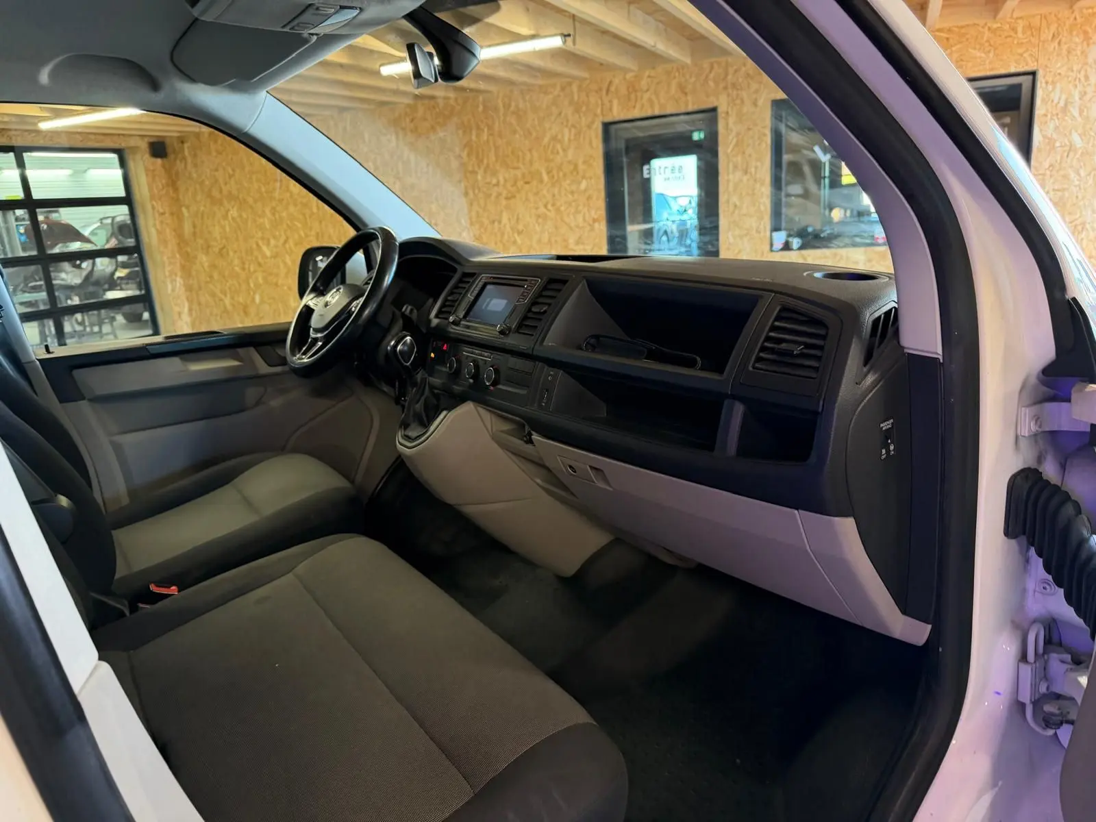 Vue intérieure côté conducteur du Volkswagen Transporter blanc 2019, tableau de bord et sièges en tissu noir visibles.