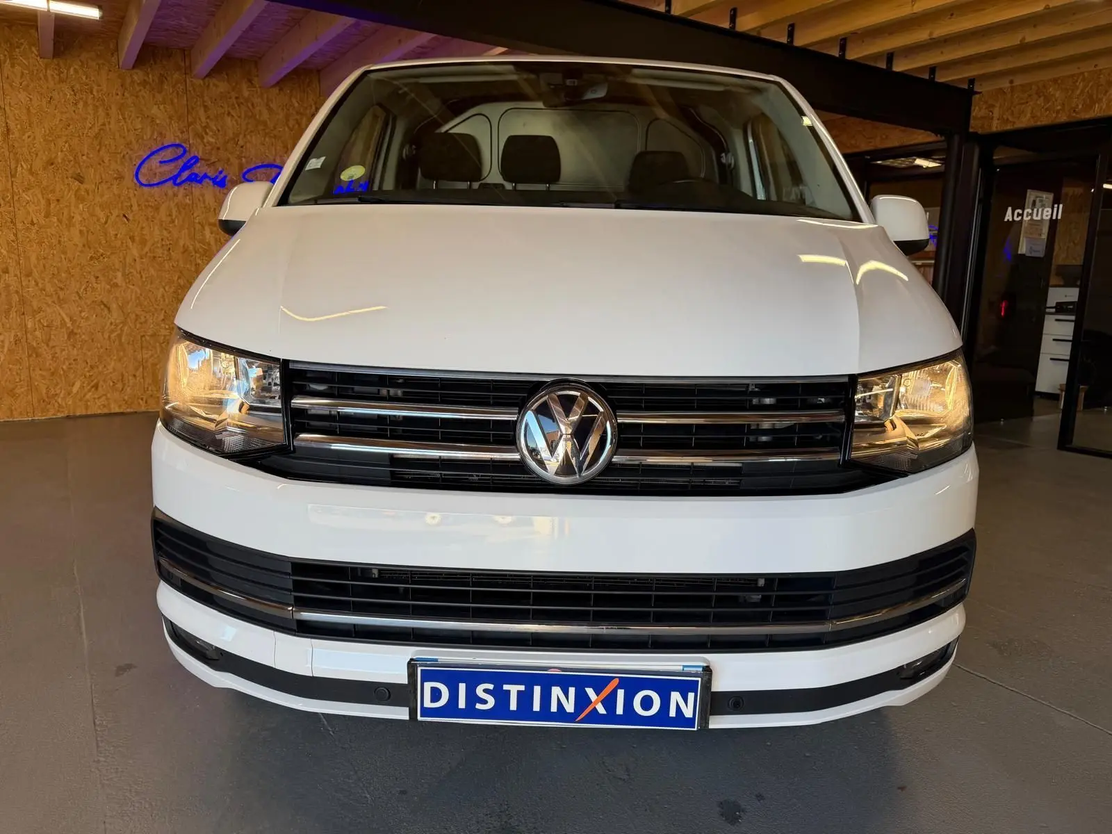 Vue frontale d’un Volkswagen Transporter blanc 2019 avec calandre noire et phares allumés en intérieur showroom.