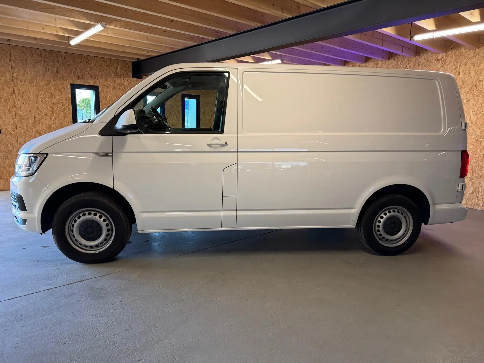 Vue de profil côté gauche d’un utilitaire Volkswagen Transporter blanc, stationné dans un garage avec murs en bois.