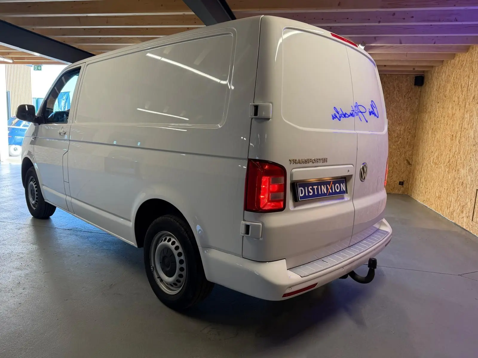 Vue 3/4 arrière droite d’un Volkswagen Transporter blanc 2019 en fourgon, avec attelage et feux arrière allumés.