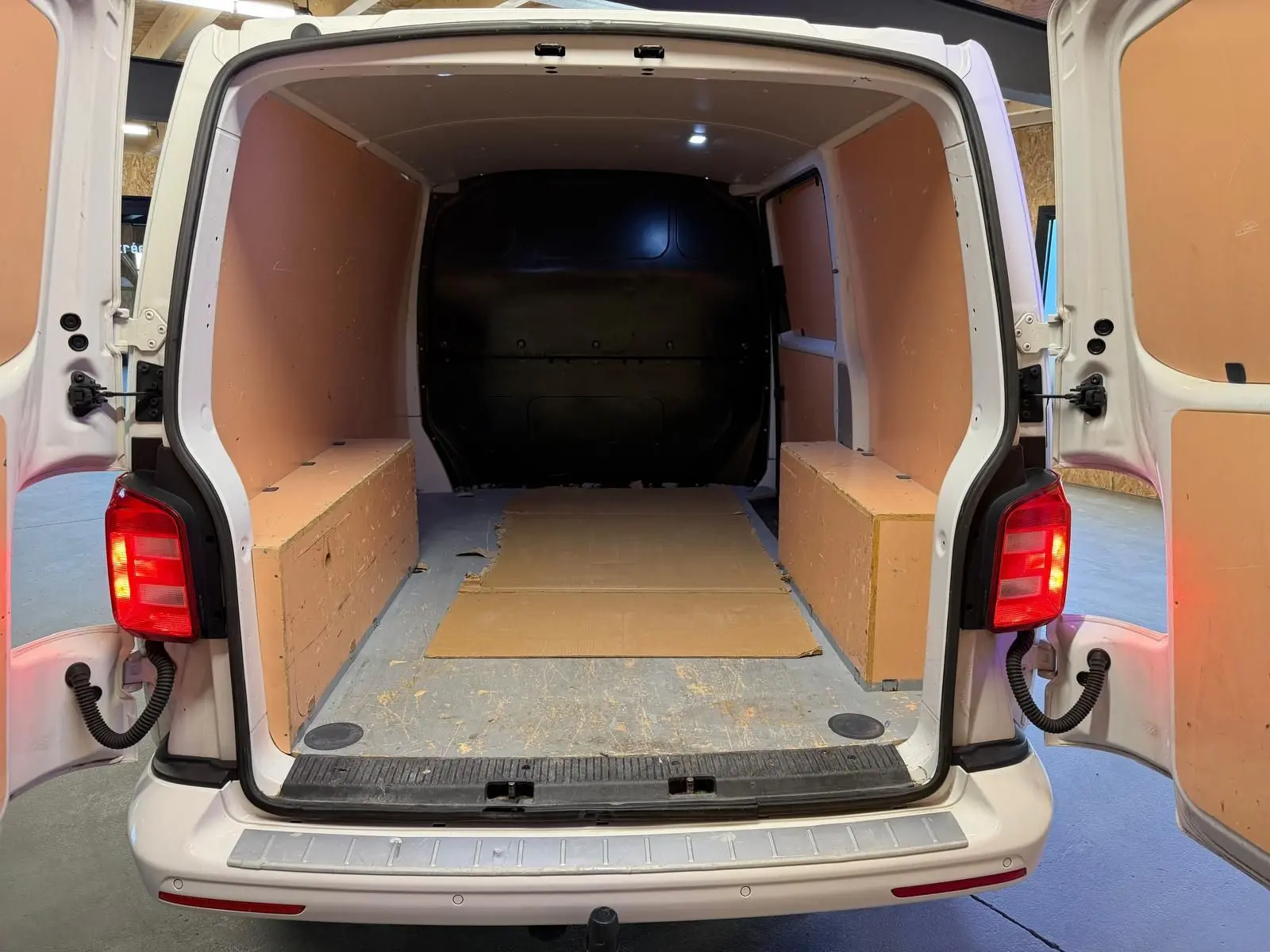 Vue arrière ouverte du Volkswagen Transporter blanc 2019, montrant l’espace de chargement vide avec panneaux latéraux en bois.