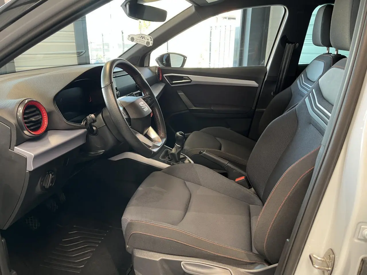 Intérieur côté conducteur du SEAT Arona blanc Nevada 2024, sièges textile noirs avec surpiqûres orange et volant sport cuir.