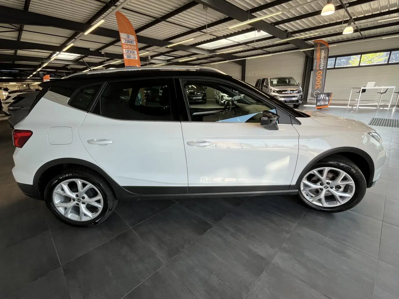 SEAT Arona blanc Nevada vue de profil côté gauche en intérieur, avec toit noir et jantes alliage 16 pouces.