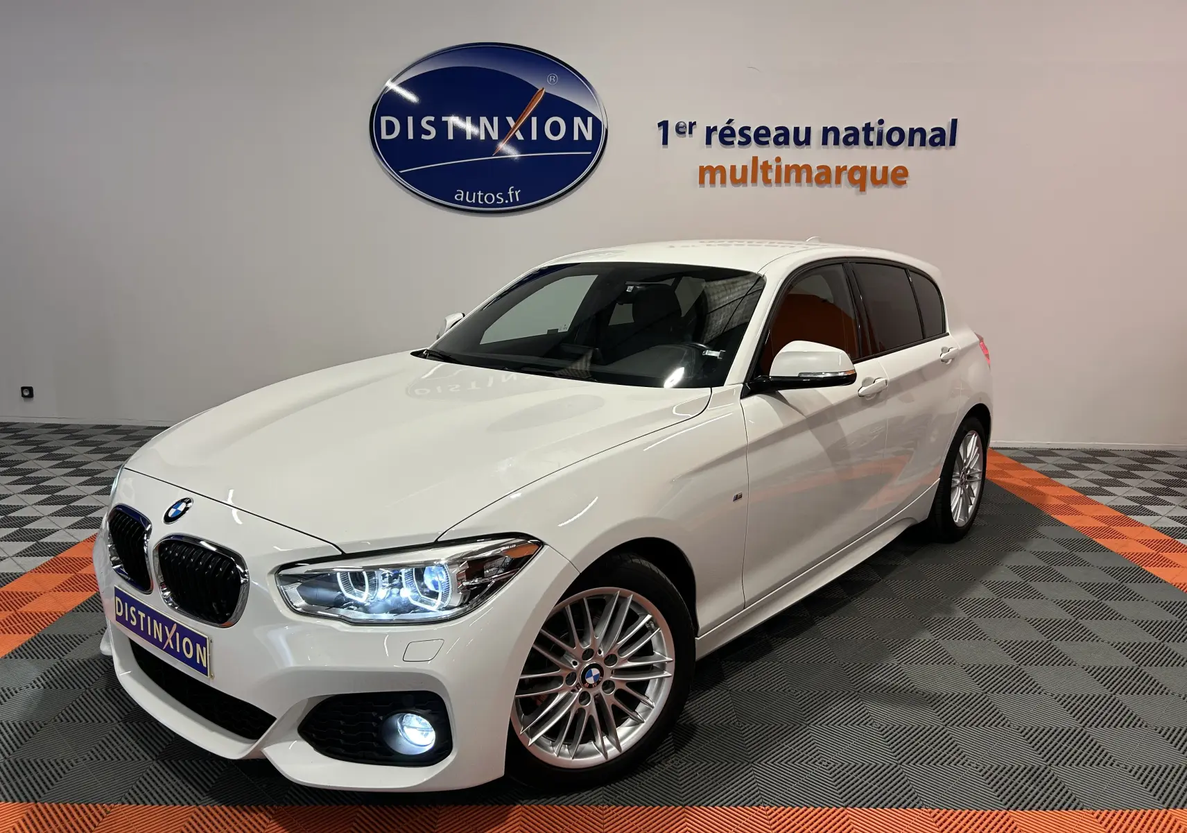 BMW Série 1 blanche vue en 3/4 avant droit, phares et feux de jour LED allumés dans un showroom.