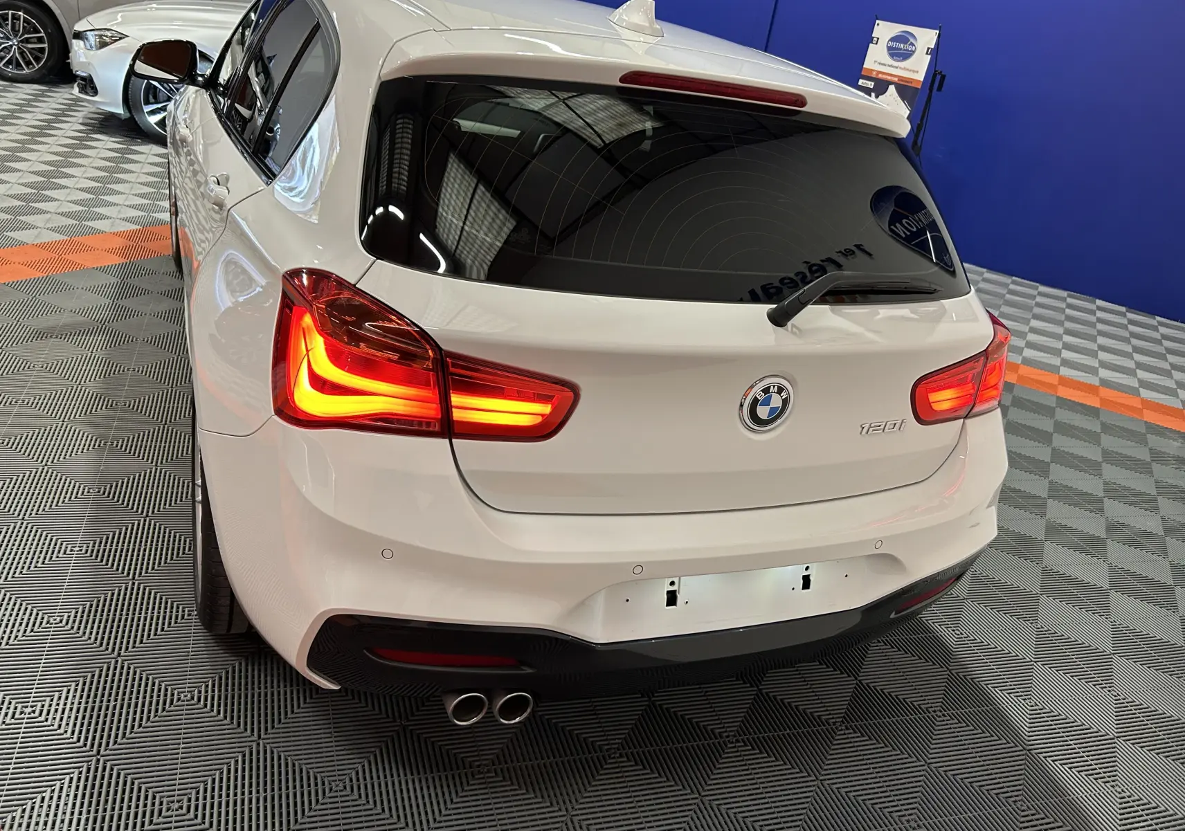 Vue 3/4 arrière d'une BMW Série 1 120iA M Sport blanche avec feux allumés et double sortie d'échappement visible